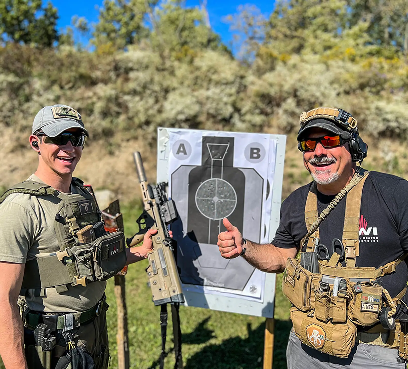 Two men in tactical gear stand outdoors, smiling proudly at a target with bullet holes during a tactical firearms training session. One holds a rifle, while the other gives a thumbs-up. Bushes and trees provide the perfect backdrop to their successful day.