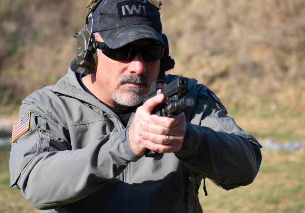 A man in grey jacket holding gun near trees.